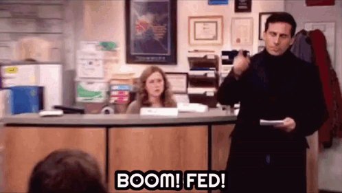 A Man Is Standing In Front Of A Desk Holding A Piece Of Paper And A Woman Is Sitting Behind Him . GIF