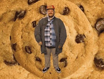 A Man Is Standing In Front Of A Large Cookie With Chocolate Chips GIF