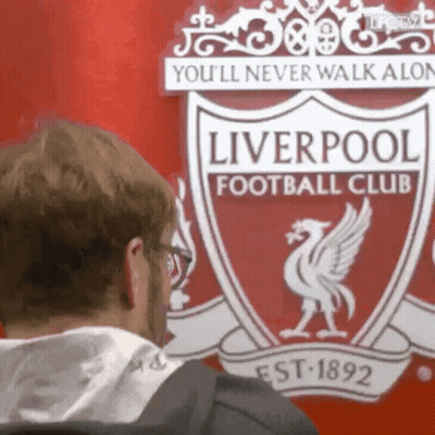 A Man Is Standing In Front Of A Liverpool Football Club Logo . GIF