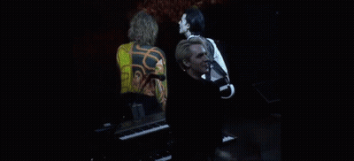 A Man Is Standing In Front Of A Piano On A Stage While A Woman Stands Behind Him . GIF