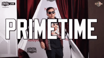 A Man Is Standing In Front Of A Red Curtain With The Words Primetime Written Above Him GIF