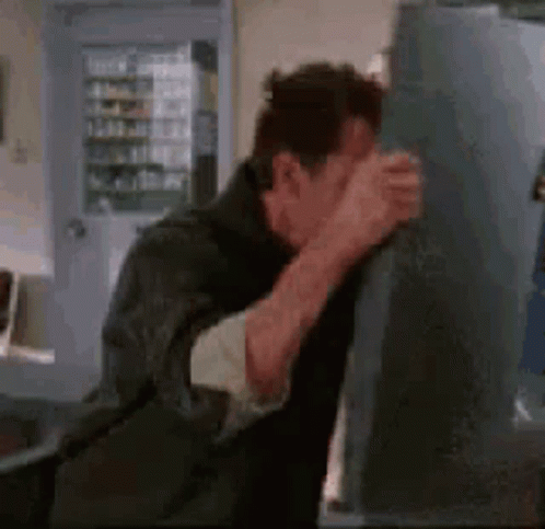 A Man Is Standing In Front Of A Refrigerator Covering His Face . GIF
