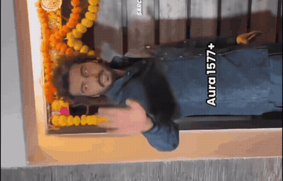 A Man Is Standing In Front Of A Wall With Flowers And A Sign That Says Aura 1577+ On It GIF