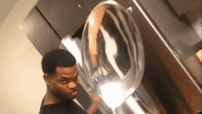 A Man Is Standing In Front Of A Washing Machine And Looking At The Reflection Of Himself In The Mirror . GIF