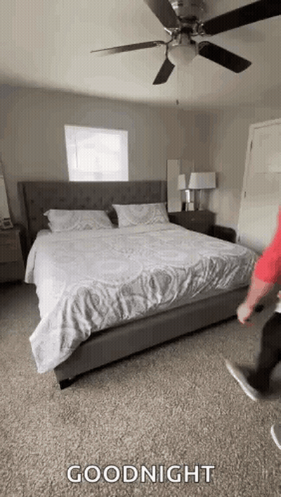 A Man Is Standing Next To A Bed In A Bedroom With A Ceiling Fan And A Window . GIF