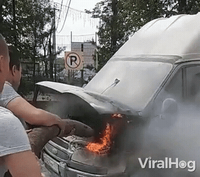A Man Is Standing Next To A Burning Van With Smoke Coming Out Of It . GIF