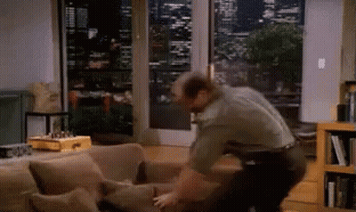 A Man Is Standing Next To A Couch In A Living Room Looking Out A Window GIF
