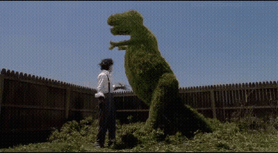 A Man Is Standing Next To A Dinosaur Made Of Bushes . GIF