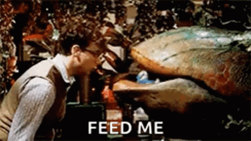 A Man Is Standing Next To A Monster That Says `` Feed Me '' On The Screen . GIF