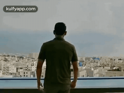 A Man Is Standing On A Balcony Overlooking A City And A Swimming Pool . GIF