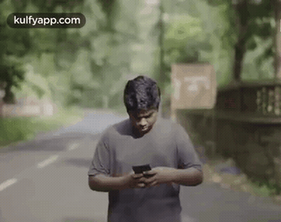 A Man Is Standing On A Road Looking At His Phone . GIF