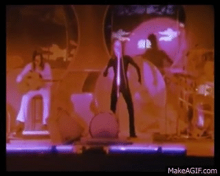 A Man Is Standing On A Stage In Front Of A Drum Set While A Woman Plays A Guitar . GIF