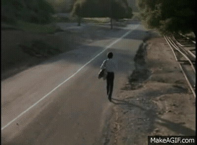 A Man Is Standing On The Side Of A Road Near Train Tracks . GIF