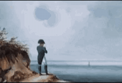 A Man Is Standing On Top Of A Cliff Overlooking The Ocean Looking Through Binoculars . GIF