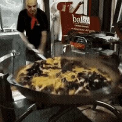 A Man Is Stirring A Large Pan Of Food In Front Of A Sign That Says Bake GIF