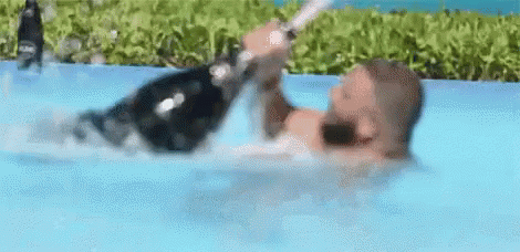 A Man Is Swimming In A Pool Holding A Bottle Of Champagne . GIF
