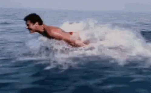 A Man Is Swimming In The Ocean With A Shark Behind Him GIF