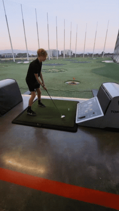 A Man Is Swinging A Golf Club At A Golf Ball On A Mat That Says Top GIF