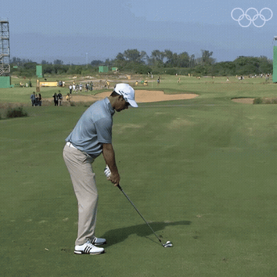 A Man Is Swinging A Golf Club On A Golf Course With The Olympic Rings In The Background GIF