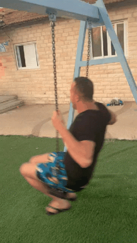 A Man Is Swinging On A Blue Swing Set In Front Of A Brick Building GIF