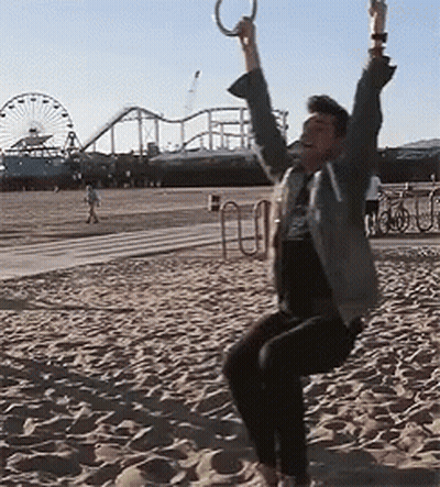 A Man Is Swinging On A Rope Swing On A Beach With A Roller Coaster In The Background GIF