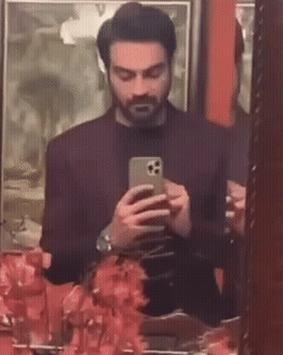 A Man Is Taking A Selfie In Front Of A Mirror While Looking At His Phone . GIF