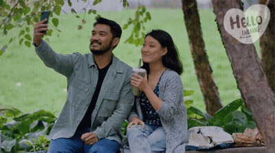 A Man Is Taking A Selfie With A Woman While They Sit Under A Tree With A Hello Again Sign Behind Them GIF