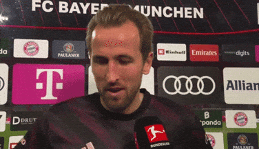 A Man Is Talking Into A Microphone In Front Of A Fc Bayern Munchen Banner GIF