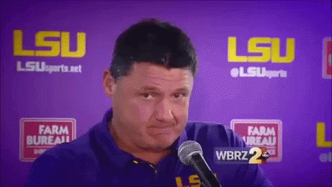 a man is talking into a microphone in front of a purple wall with lsu written on it