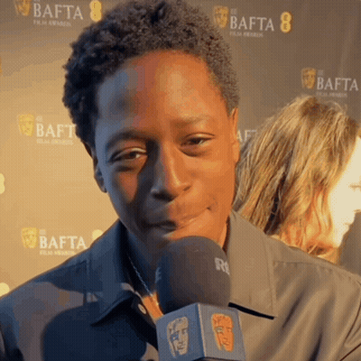 A Man Is Talking Into A Microphone In Front Of A Wall That Says Bafta GIF