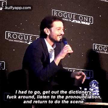A Man Is Talking Into A Microphone While Sitting At A Table In Front Of A Sign That Says Rogue One . GIF