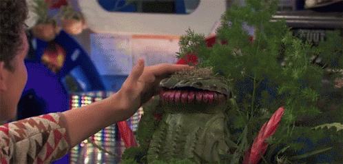 A Man Is Touching A Green Monster With Sharp Teeth Surrounded By Plants And Flowers . GIF