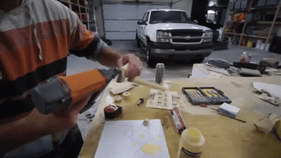 A Man Is Using A Nail Gun On A Piece Of Wood In Front Of A Truck GIF