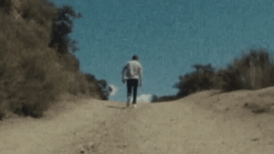 A Man Is Walking Down A Dirt Road In The Woods . GIF