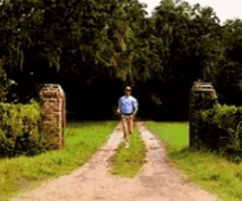 A Man Is Walking Down A Dirt Road Surrounded By Trees GIF