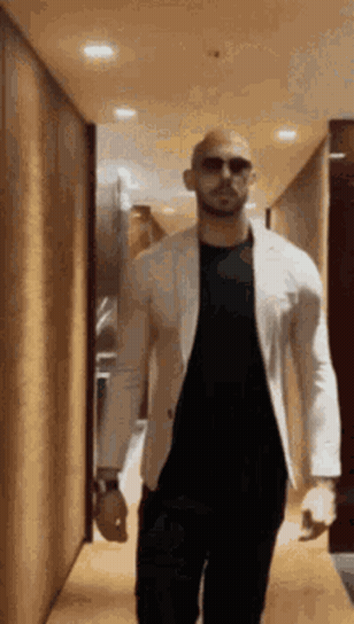A Man Is Walking Down A Hallway Wearing Sunglasses And A White Jacket . GIF