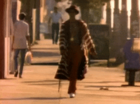 A Man Is Walking Down A Sidewalk Wearing A Hat And A Coat . GIF