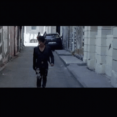 A Man Is Walking Down A Street With A Skateboard In His Hand GIF