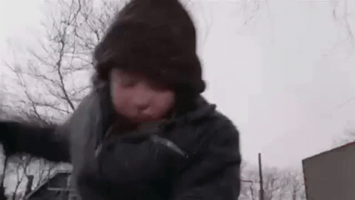 A Man Is Wearing A Hooded Jacket And Gloves While Standing In The Snow . GIF