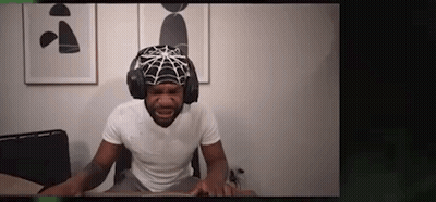 A Man Is Wearing A Spider Web Hat And Headphones While Sitting At A Desk . GIF