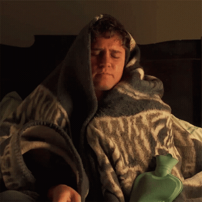 A Man Is Wrapped In A Blanket With A Green Hot Water Bottle GIF