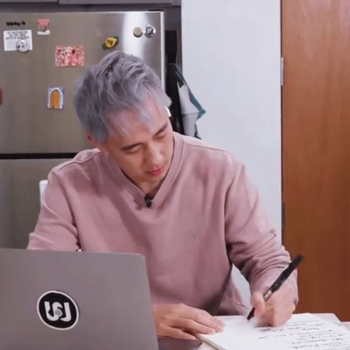 A Man Is Writing On A Piece Of Paper In Front Of A Laptop With A Sticker On It That Says Lj GIF