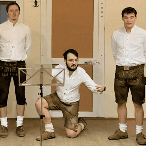 A Man Kneeling Down In Front Of A Music Stand With Two Other Men Standing Behind Him GIF