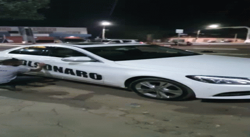 A Man Kneeling Next To A White Car That Says Bolsonaro On The Side GIF