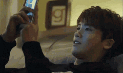 A Man Laying On A Bed Looking At A Cell Phone With The Letter G On The Wall Behind Him GIF