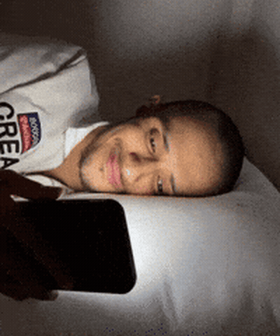 A Man Laying On A Bed Looking At His Phone With A Shirt That Says Brooklyn Great GIF