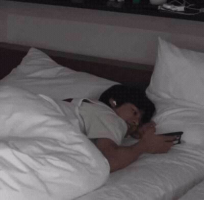 A Man Laying On A Bed With White Sheets Looking At His Phone GIF