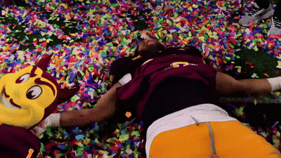 A Man Laying On A Pile Of Confetti With A Mascot In The Background GIF