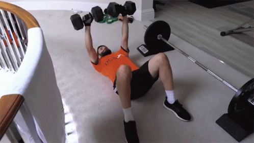 A Man Laying On The Floor Lifting Dumbbells With A Shirt That Says Ucla GIF