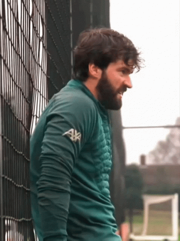 A Man Leaning Against A Fence Wearing A Green Sweater With Axa On The Sleeves GIF
